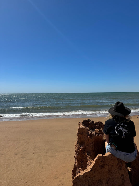 A Lady sitting on red rocks of the Pilbara with the Unearth Style Co Horizon Palms shirt looking out at the beautiful ocean.