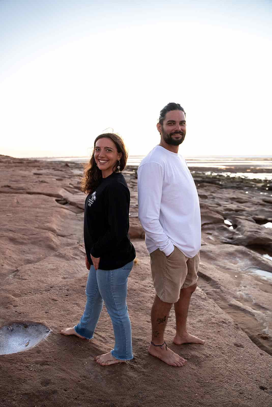 A couple standing on the rocks near the beach looking happy in the camera wearing Unearth Style Co long sleeve shirts